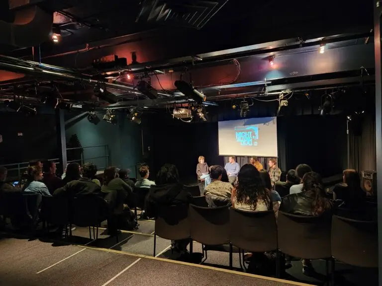 The basement theatre space and the Palmerston Public Library, seats full of audience members, and four speaks on the stage.